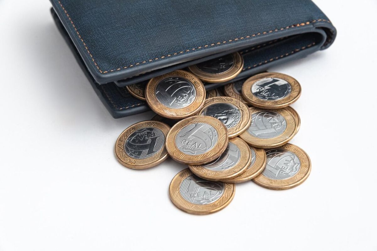 Uma carteira preta de onde está saindo moedas de R$ 1,00