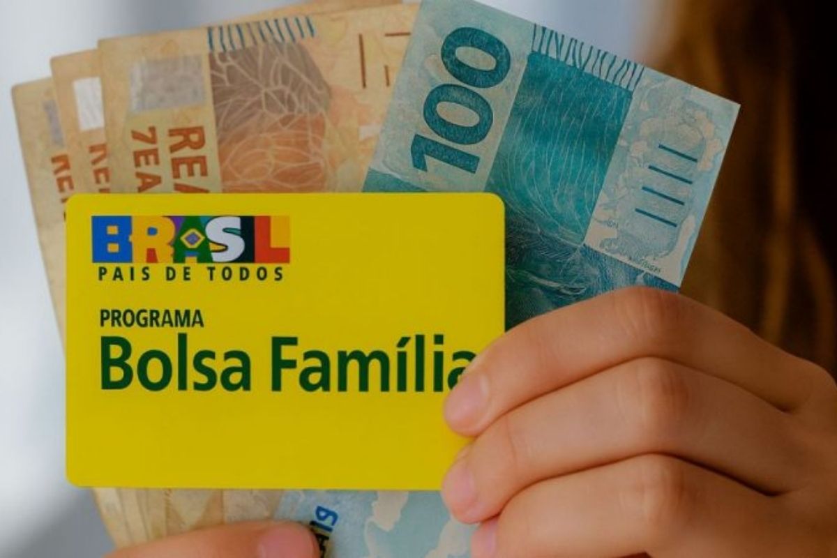 Cartão do Bolsa Família na mão de uma pessoa com notas de R$ 100,00 e R$ 50,00