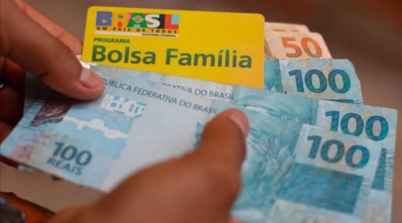 Cartão do Bolsa Família na mão de uma pessoa com notas de R$ 100,00 e R$ 50,00
